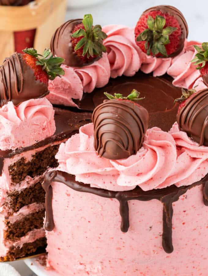 Close-up of a chocolate strawberry cake with pink strawberry buttercream, chocolate ganache drip, and chocolate-covered strawberries on top.
