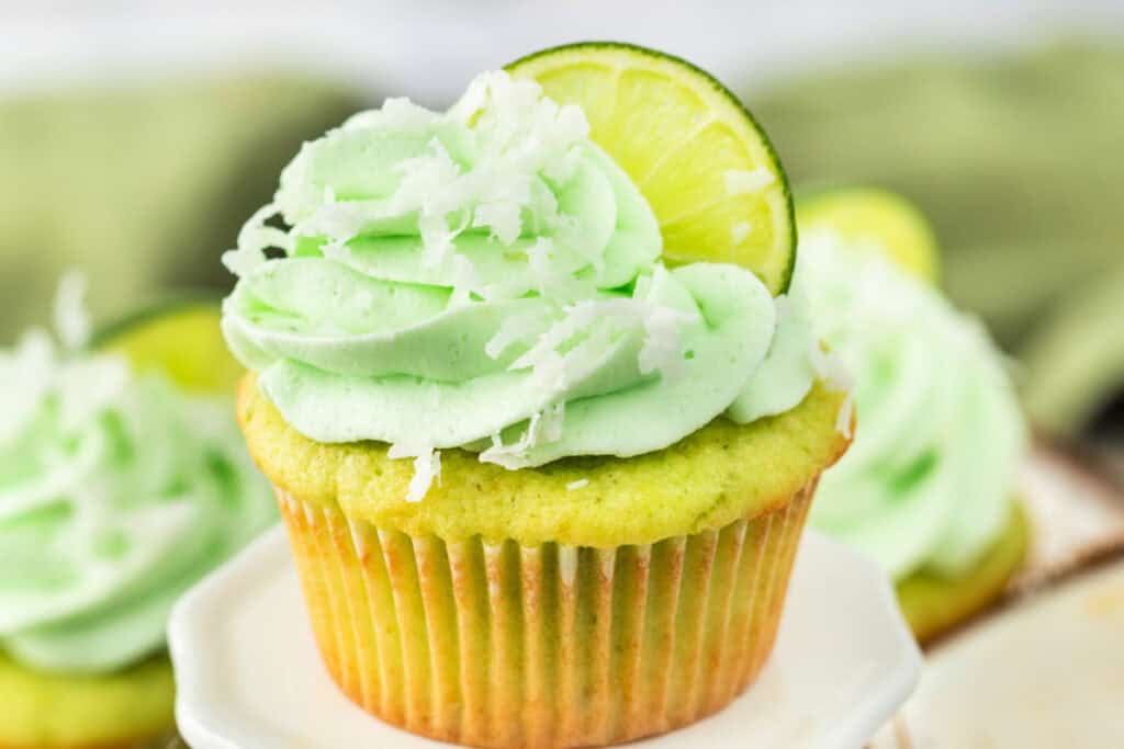 Frosted coconut lime cupcake on a white stand with more cupcakes in background