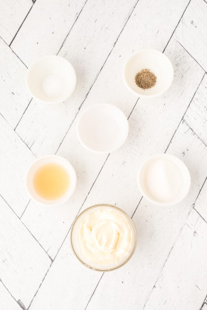 Small bowls of individual coleslaw dressing ingredients on a white herringbone surface.