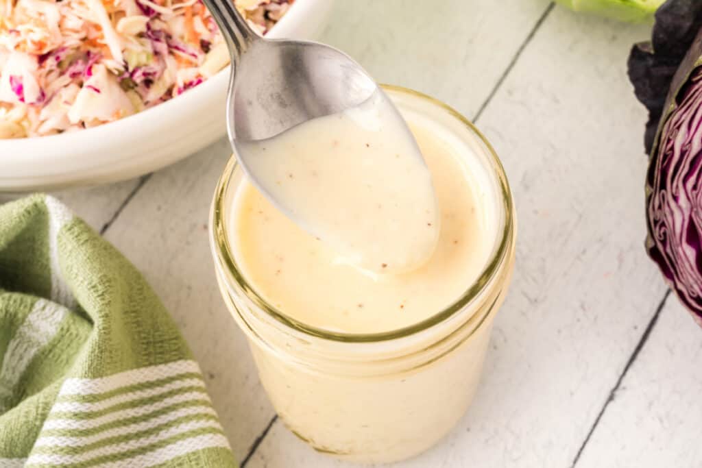 Close-up of creamy coleslaw dressing on a spoon over a glass jar, with coleslaw in the background.