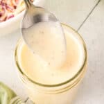 Overhead view of a spoon lifting creamy coleslaw dressing from a jar with a green towel nearby.