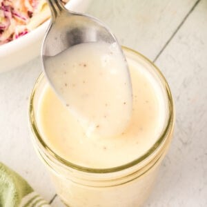 Overhead view of a spoon lifting creamy coleslaw dressing from a jar with a green towel nearby.