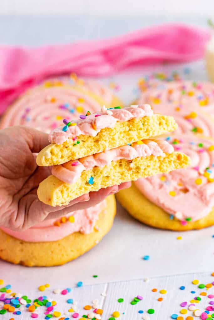 Hand holding a frosted confetti cookie broken in half, showing soft and fluffy texture inside.
