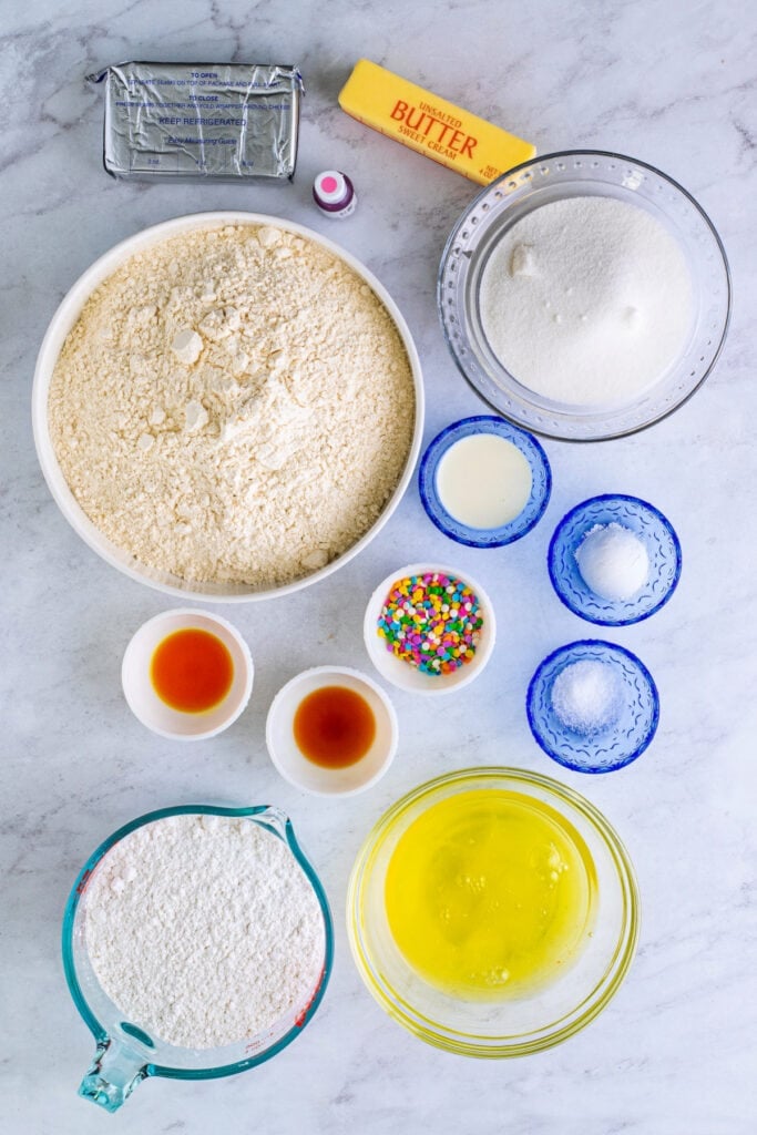 Overhead view of cookie dough ingredients, including butter, sugar, flour, egg whites, sprinkles, and extracts on a marble background.