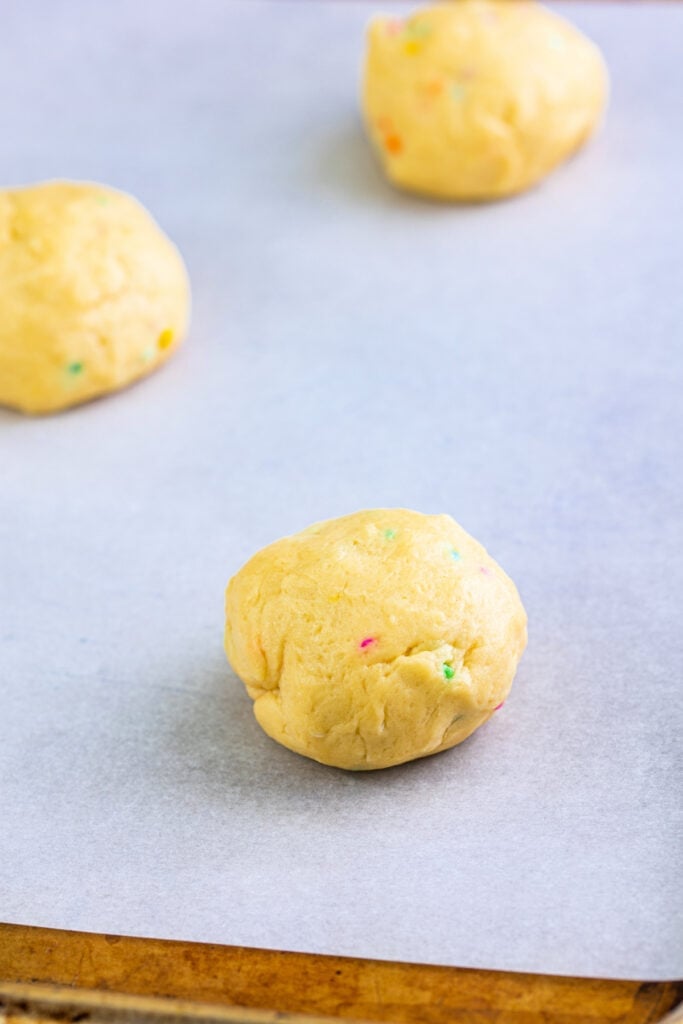Close-up of cookie dough ball with colorful sprinkles on parchment-lined baking sheet.
