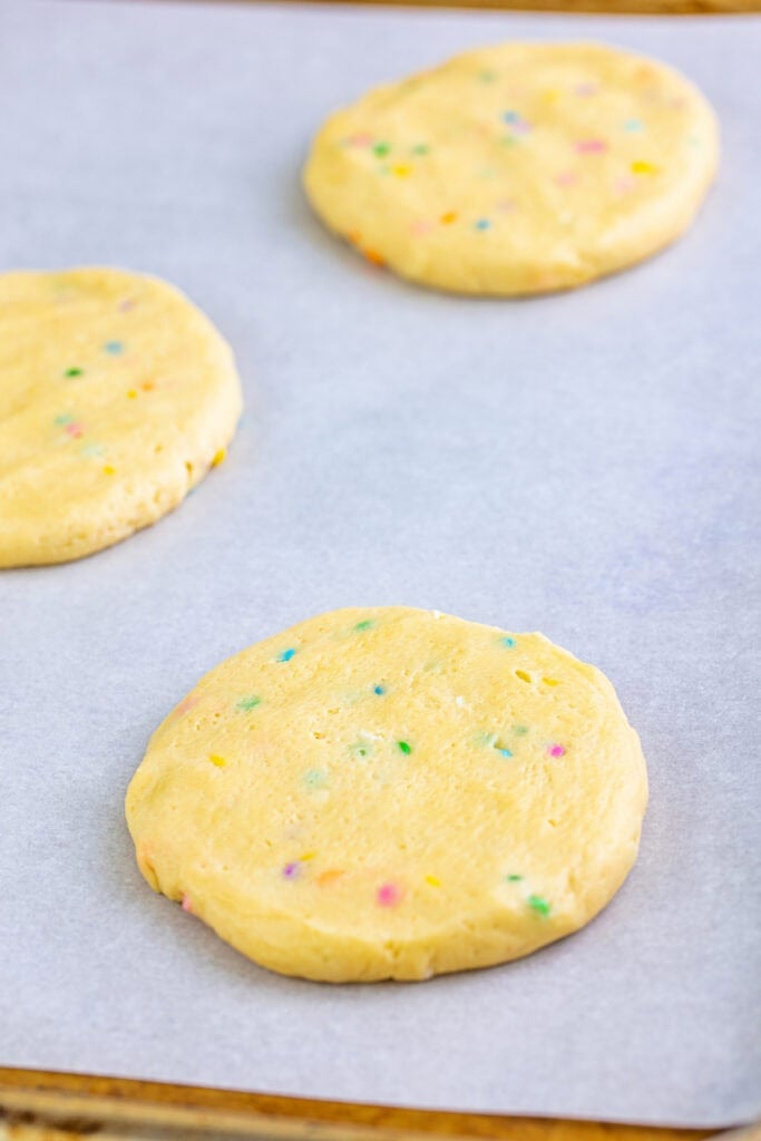 Flattened cookie dough circles with visible sprinkles ready to bake on a parchment-lined baking sheet.