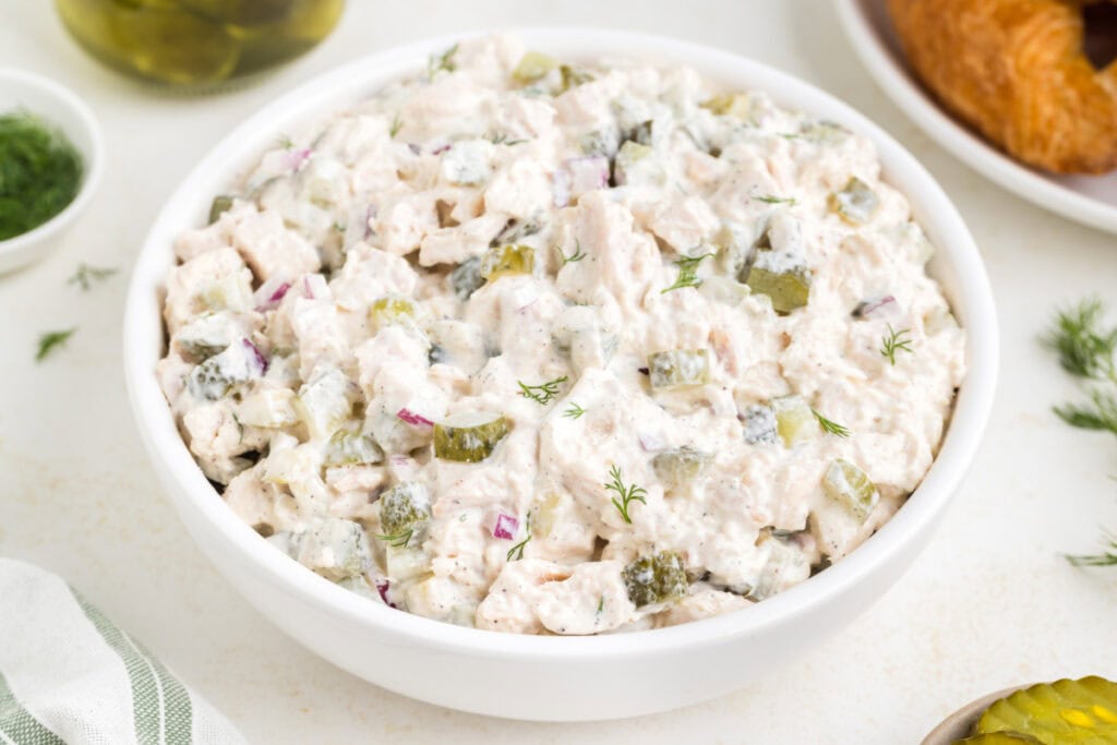 Finished chicken salad served in a white bowl, surrounded by pickles, croissants, and fresh dill.
