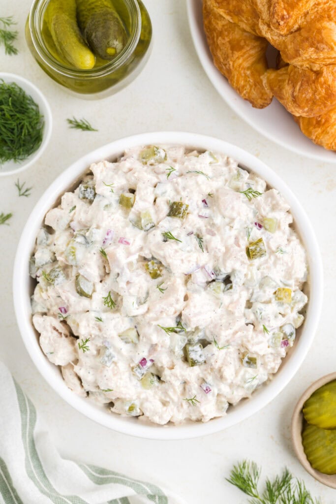 Overhead view of dill pickle chicken salad in a white bowl, surrounded by croissants, pickles, and fresh dill.