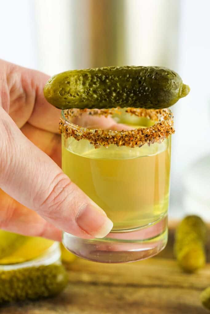 Hand holding a shot glass rimmed with tajin and topped with a gherkin pickle