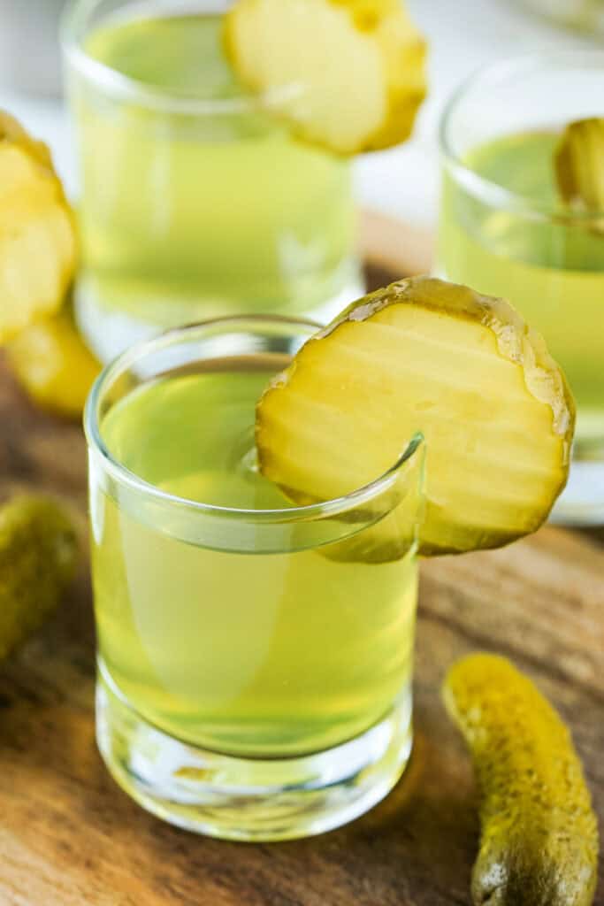 Angled view of multiple pickle shots with pickle chip garnishes on a wooden board