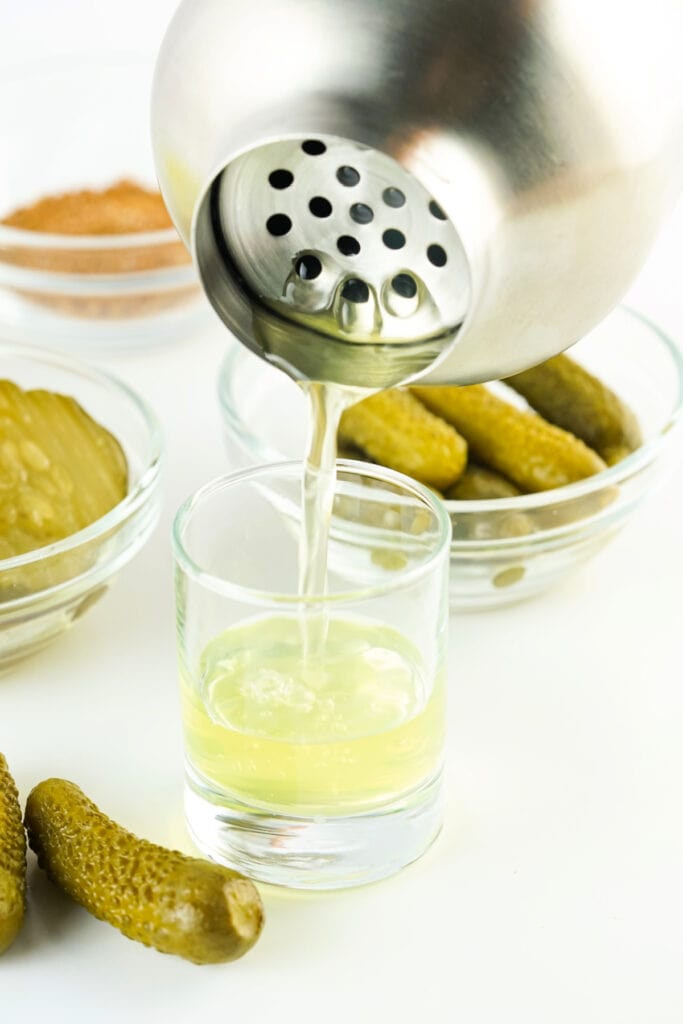 Cocktail shaker pouring pickle juice mixture into a shot glass with pickles in bowls