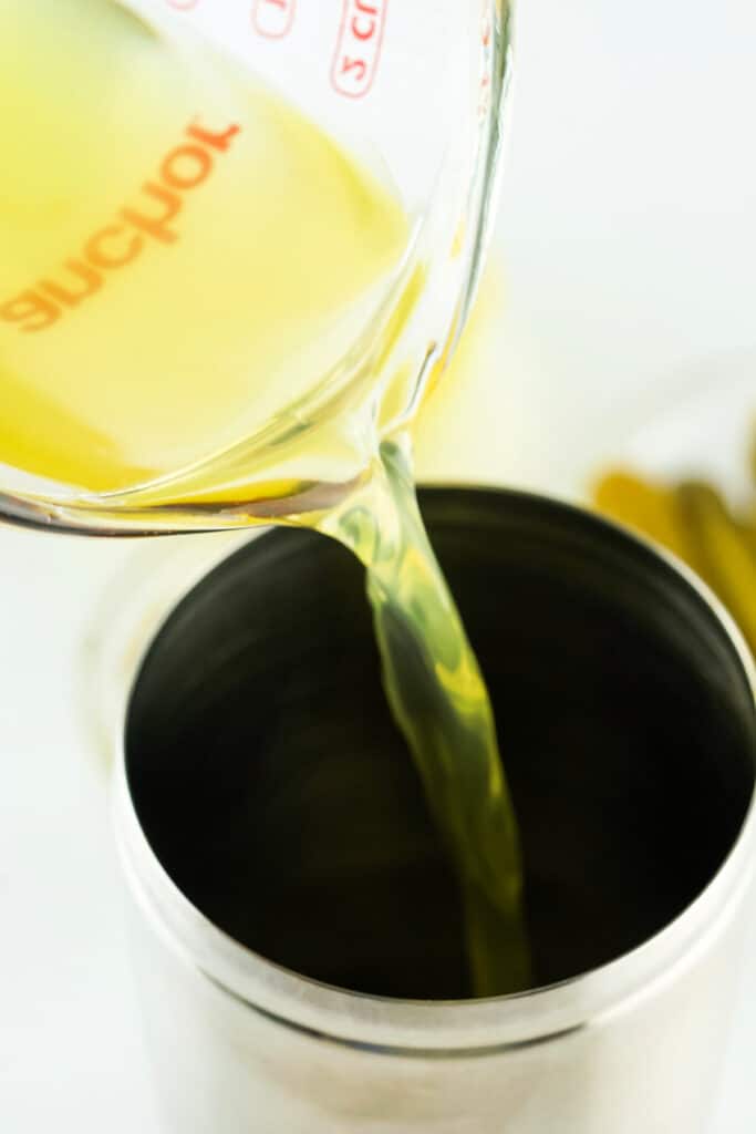 Measuring cup pouring pickle juice into a stainless steel cocktail shaker