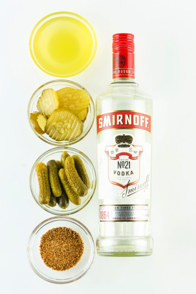 Flat lay of vodka bottle, pickle juice, tajin, and pickles in small bowls