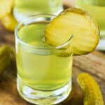 Square format close-up of a dill pickle shot with pickle chip garnish on a wood board