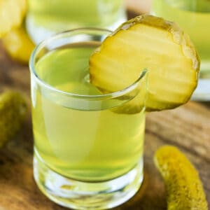 Square format close-up of a dill pickle shot with pickle chip garnish on a wood board