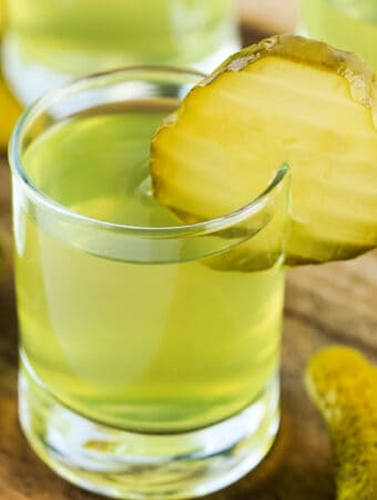 Square format close-up of a dill pickle shot with pickle chip garnish on a wood board