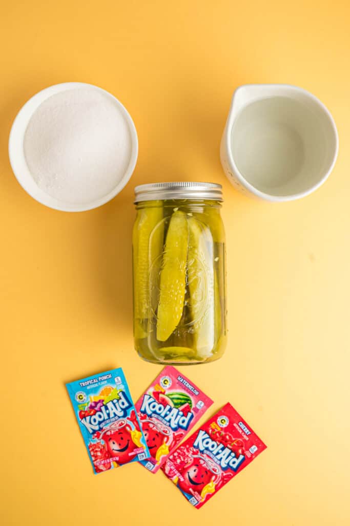 	Ingredients for Kool-Aid pickles laid out on a yellow background, including sugar, water, pickle jar, and Kool-Aid packets.