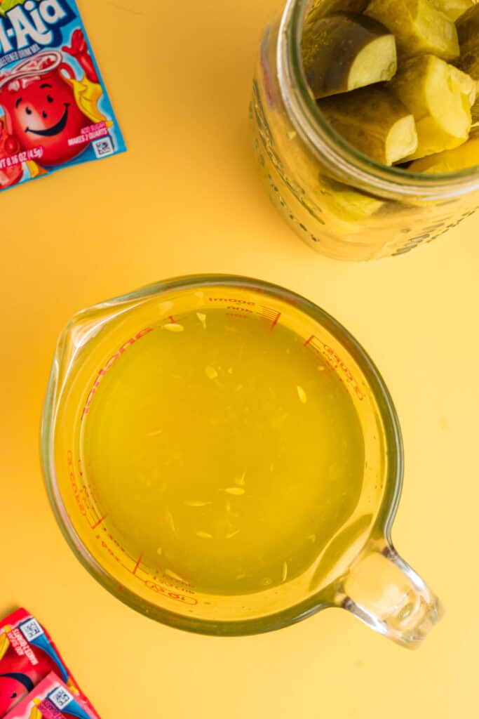 Pickle brine poured into a measuring cup next to pickles and Kool-Aid packets.