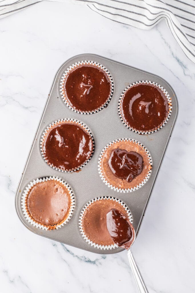 Spoon spreading ganache onto baked mini chocolate cheesecakes in a muffin pan