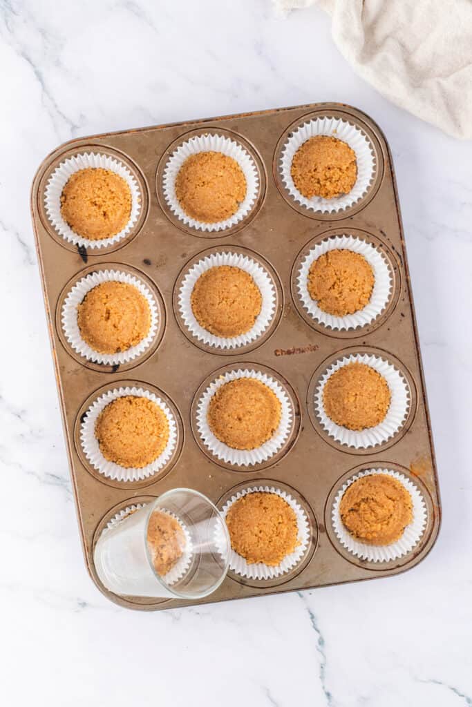 Muffin pan lined with white cupcake liners, each filled with pressed graham cracker crust. A small glass is shown pressing the crust into shape.