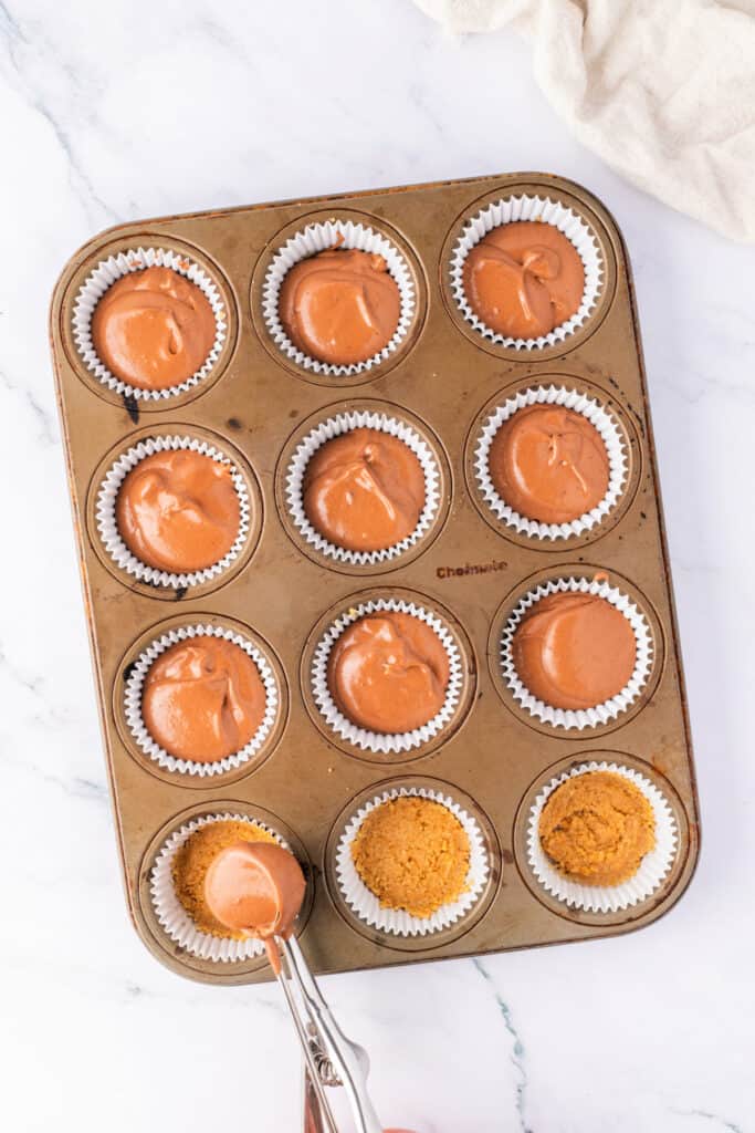 Chocolate cheesecake filling being spooned over graham cracker crust in a lined muffin pan. Some cups are already filled.