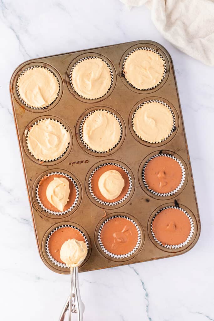 Peanut butter cheesecake layer being added over the chocolate layer in a lined muffin pan using a cookie scoop.