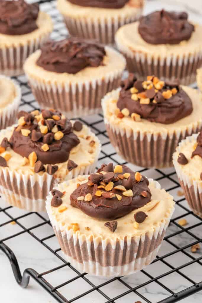 Finished mini cheesecakes topped with chocolate ganache, chopped peanuts, and mini chocolate chips cooling on a wire rack.