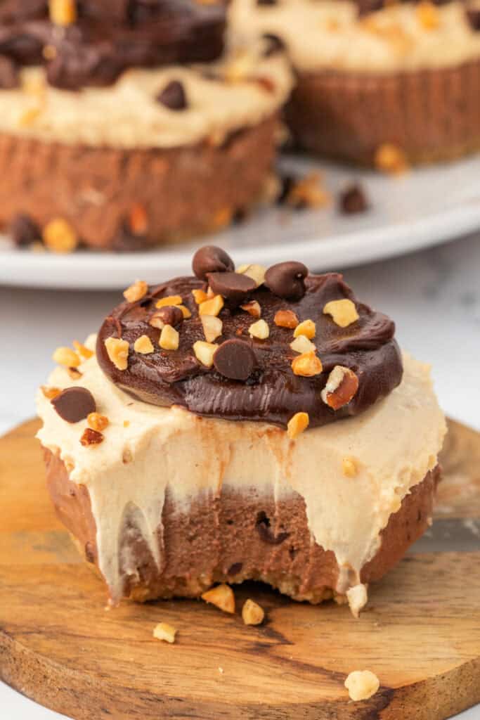 Close-up of a mini cheesecake with a bite taken out, showing chocolate and peanut butter layers, topped with ganache and chopped peanuts.