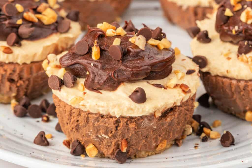 Close-up of a mini cheesecake topped with a generous swirl of chocolate ganache and sprinkled with peanuts and mini chocolate chips.
