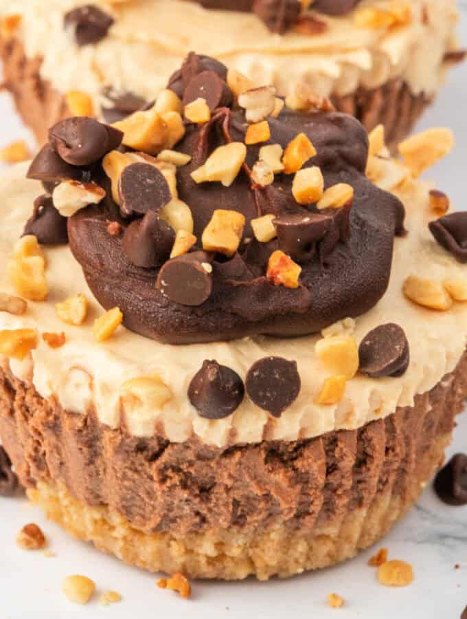 Overhead view of mini cheesecakes on a white plate, decorated with chocolate ganache, mini chocolate chips, and chopped peanuts.