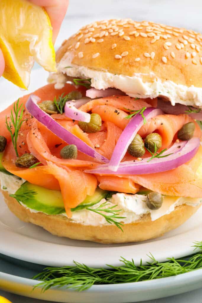 Close-up of a sesame bagel sandwich being finished with a squeeze of lemon over smoked salmon, red onions, and capers.