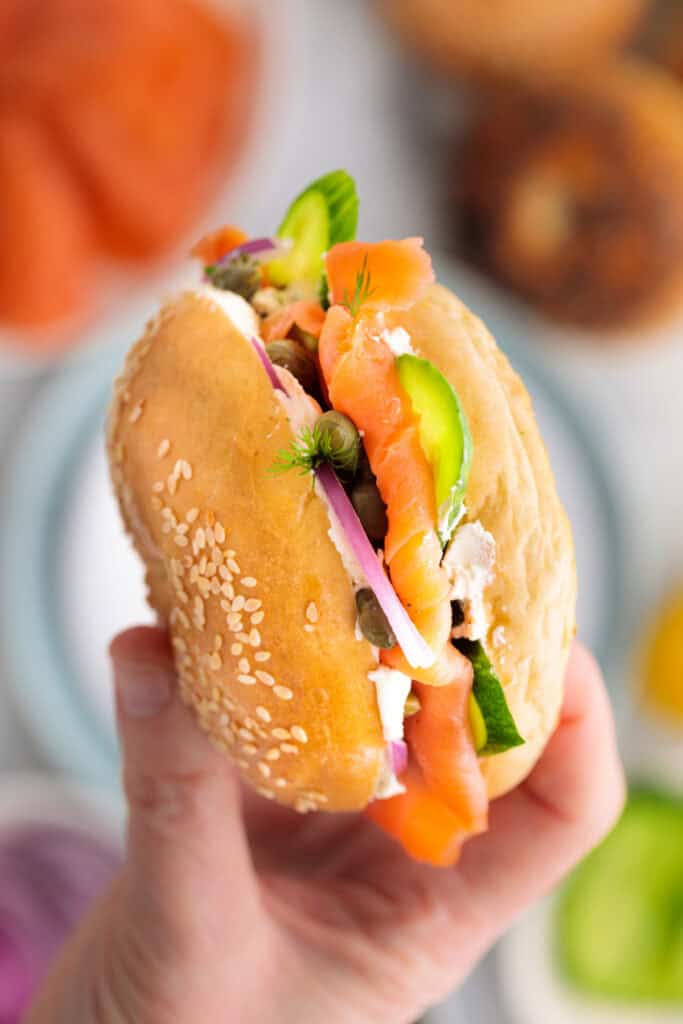 Hand holding a fully assembled smoked salmon bagel with sesame seed bun, cream cheese, capers, cucumber, and red onion.