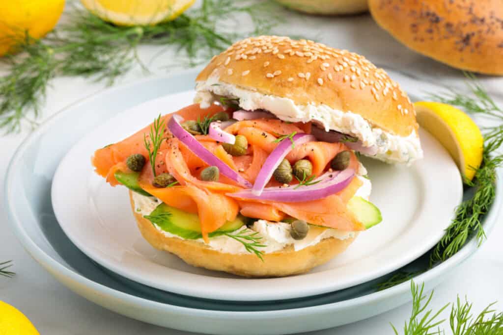 Smoked salmon bagel sandwich on a plate with fresh dill, red onion, cucumber, capers, and cream cheese, with lemon garnish in background.