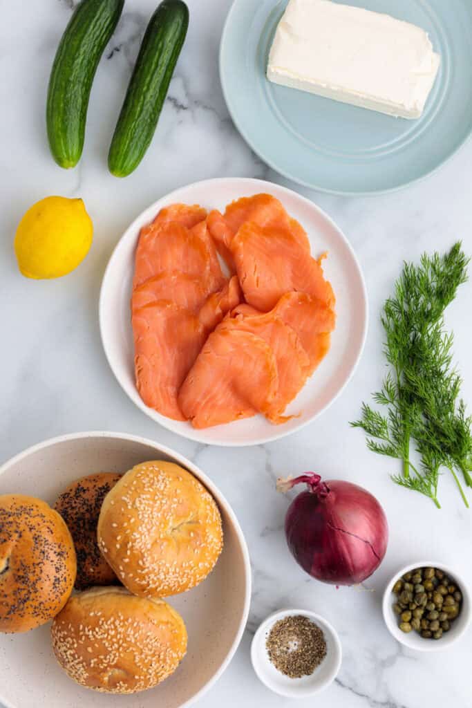 Ingredients laid out for smoked salmon bagels including bagels, cream cheese, smoked salmon, dill, cucumber, red onion, lemon, capers, and seasonings.