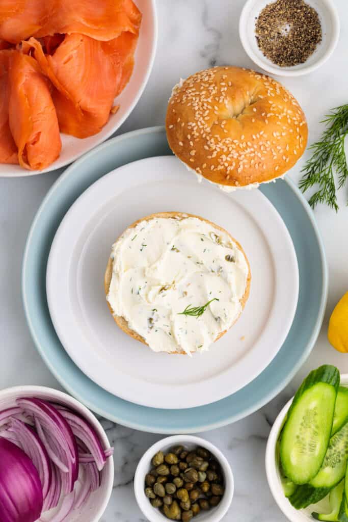 Sesame bagel half spread with herbed cream cheese surrounded by ingredients like smoked salmon, cucumber, red onion, and capers.