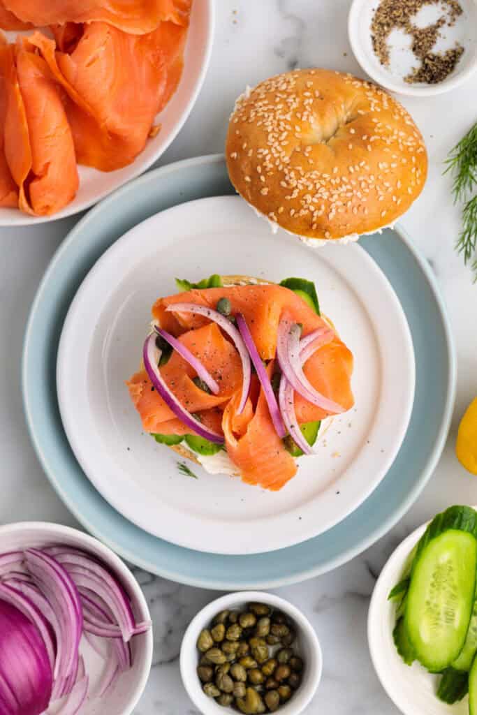 Smoked salmon bagel with red onion slices and capers added, showing a near-finished build.