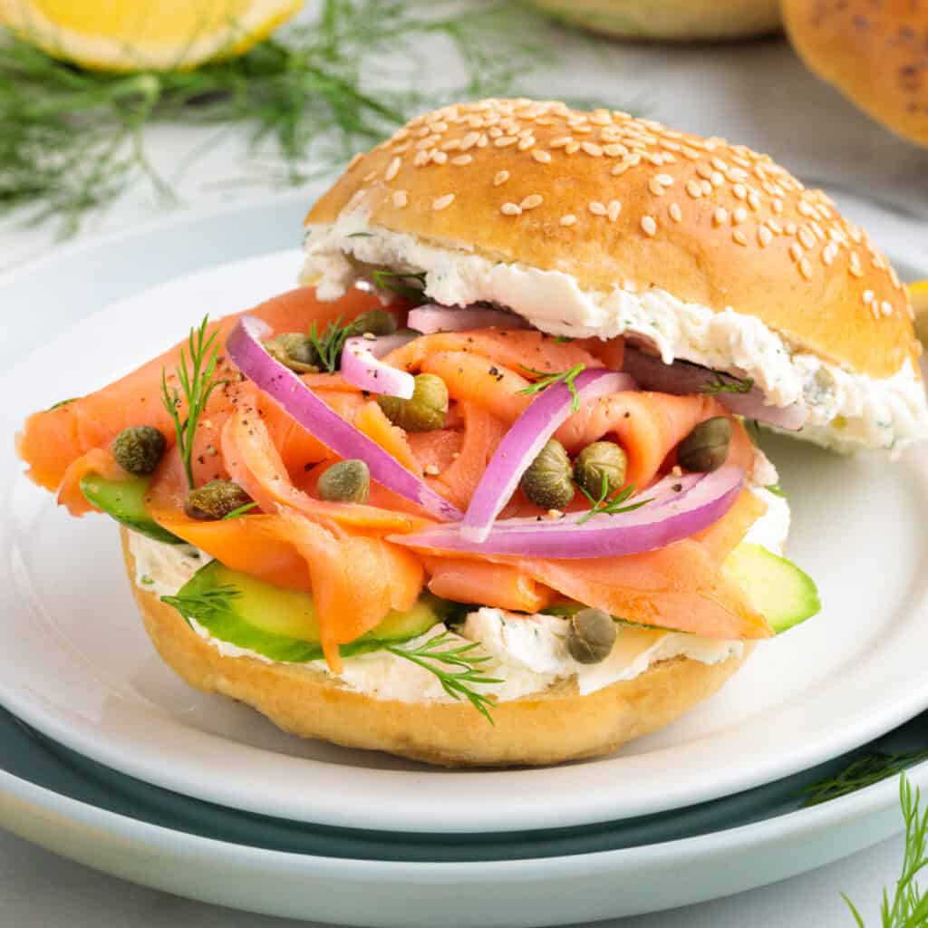 Square image of a smoked salmon bagel sandwich with cream cheese, red onions, capers, dill, and cucumber on a sesame bagel.