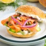 Square image of a smoked salmon bagel sandwich with cream cheese, red onions, capers, dill, and cucumber on a sesame bagel.