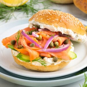 Square image of a smoked salmon bagel sandwich with cream cheese, red onions, capers, dill, and cucumber on a sesame bagel.