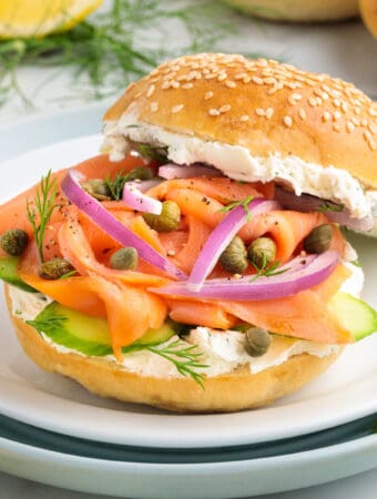 Square image of a smoked salmon bagel sandwich with cream cheese, red onions, capers, dill, and cucumber on a sesame bagel.