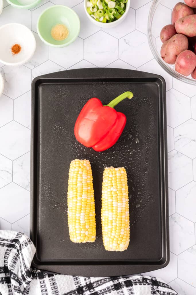 Red bell pepper and two ears of corn placed on a black baking sheet, ready to roast.