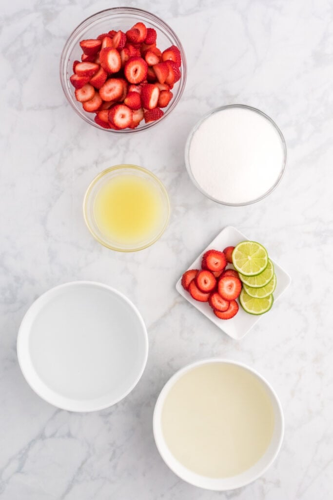 Overhead shot of fresh sliced strawberries, lime juice, sugar, coconut water, water, and lime slices arranged on a marble surface.