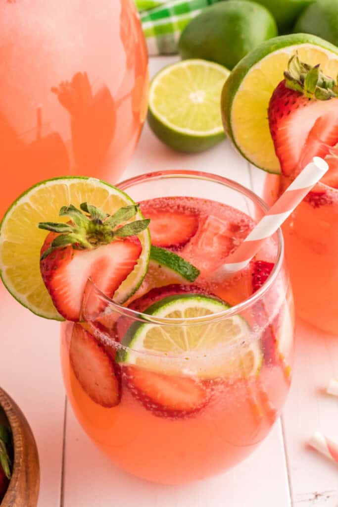 Close-up of a glass filled with strawberry limeade, garnished with fresh lime and strawberry slices, and a pink striped straw.