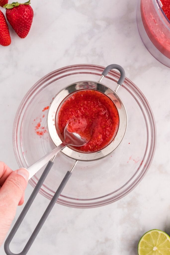 Strawberry purée being strained through a fine mesh strainer with a spoon over a glass bowl.