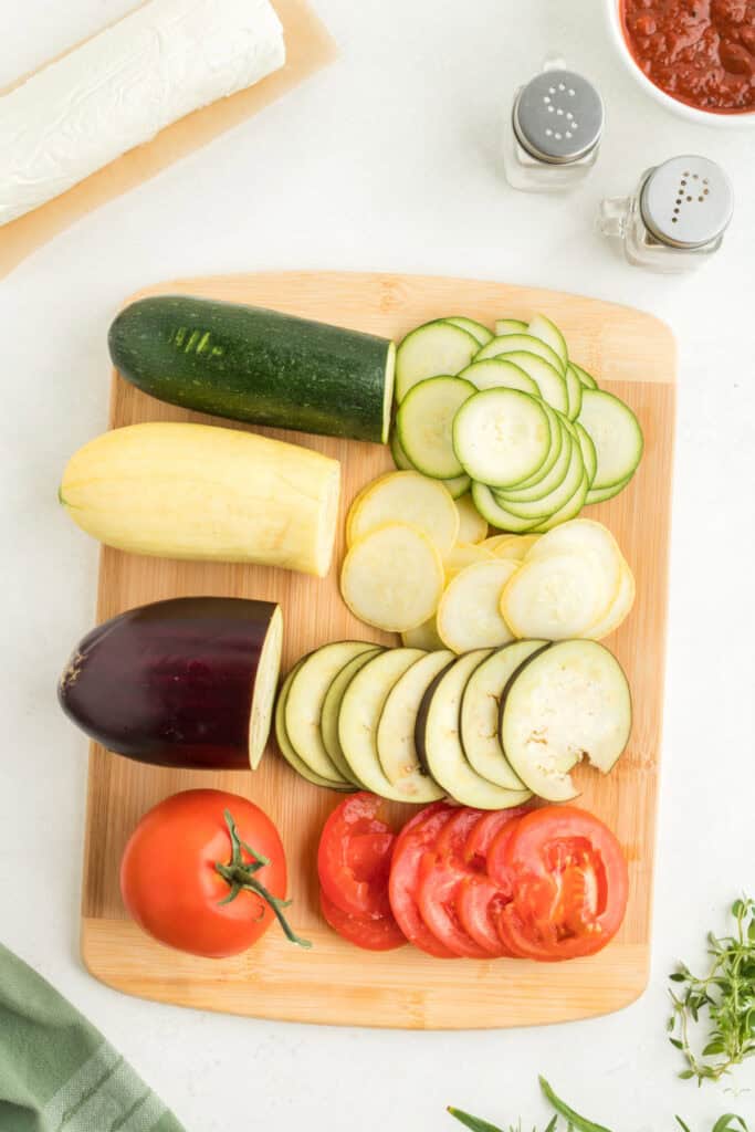 Sliced vegetables including zucchini, yellow squash, eggplant, and tomatoes arranged on a cutting board.