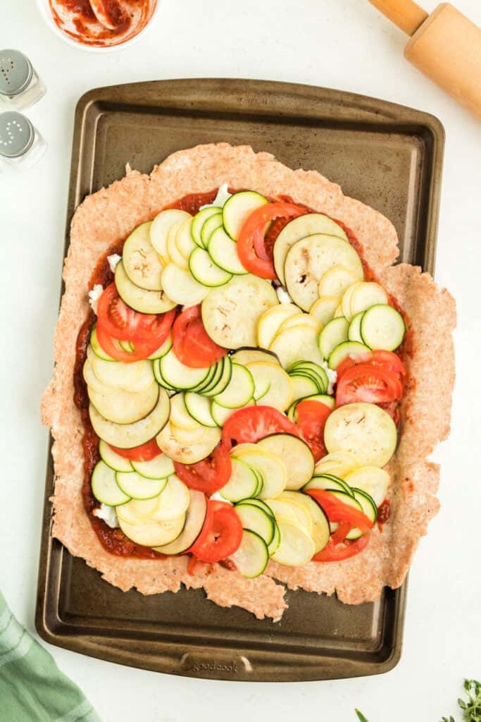Unbaked galette crust spread on a baking sheet topped with marinara, goat cheese, and layered vegetables.