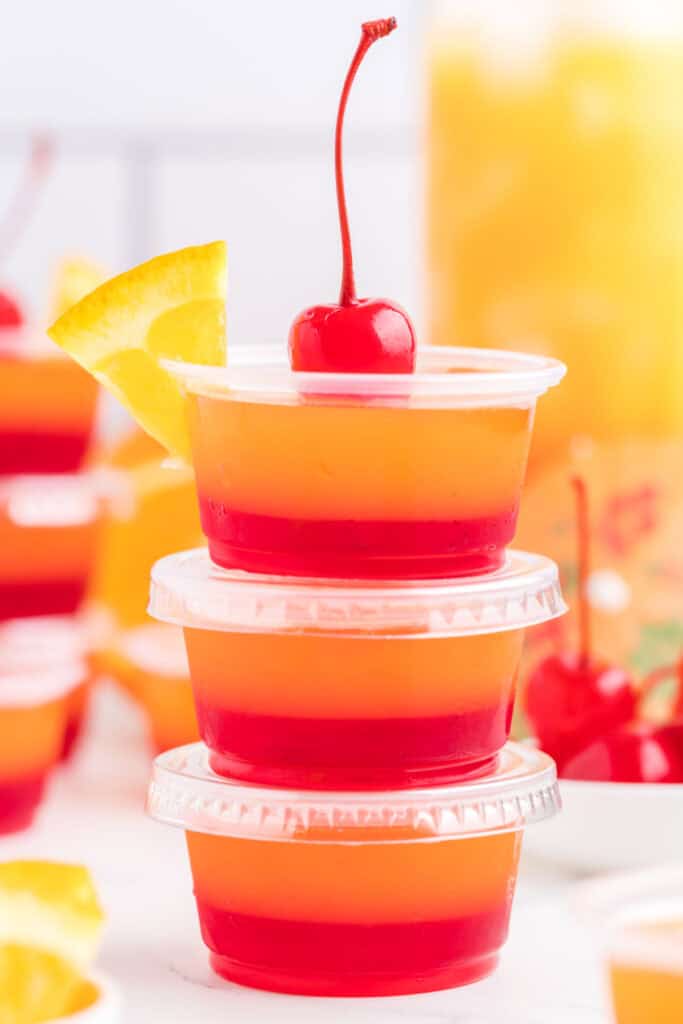 Stack of tequila sunrise jello shots with lids, topped with a maraschino cherry and orange slice.