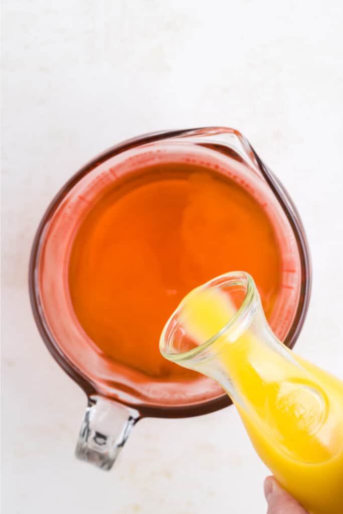 Orange juice being poured into prepared orange jello mixture in a measuring cup.