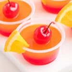 Close-up of a single tequila sunrise jello shot garnished with a maraschino cherry and orange slice.