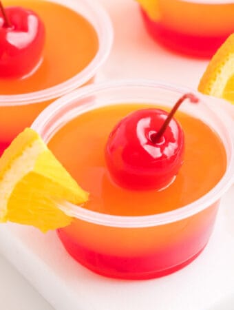 Close-up of a single tequila sunrise jello shot garnished with a maraschino cherry and orange slice.